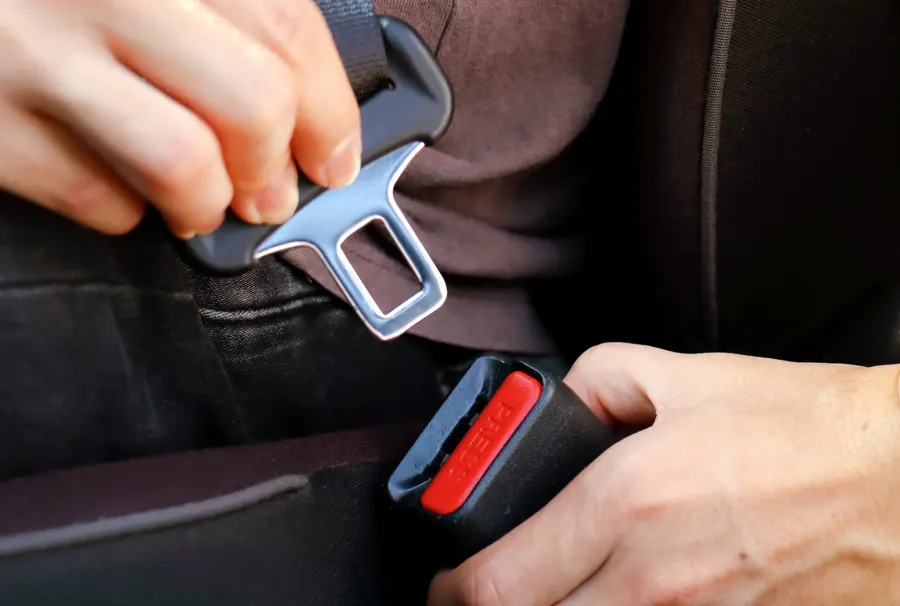 Close view of both hands inserting a seatbelt tongue plate into a buckle receiver with a red release button.